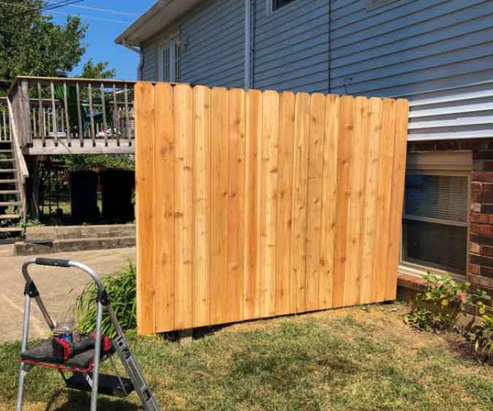 Outdoor installation of a tall wooden privacy fence panel, with tools and a ladder on a sunny day, enhancing backyard privacy.