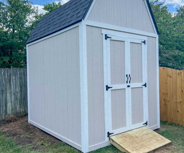 A freshly painted white outdoor storage shed with black hardware, positioned against a residential fence in a backyard setting.