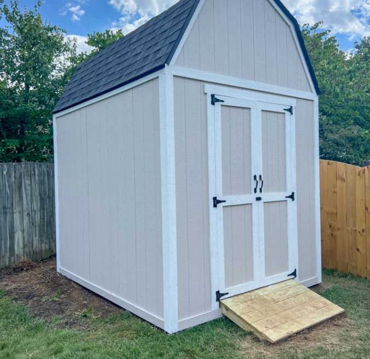 A freshly painted white outdoor storage shed with black hardware, positioned against a residential fence in a backyard setting.