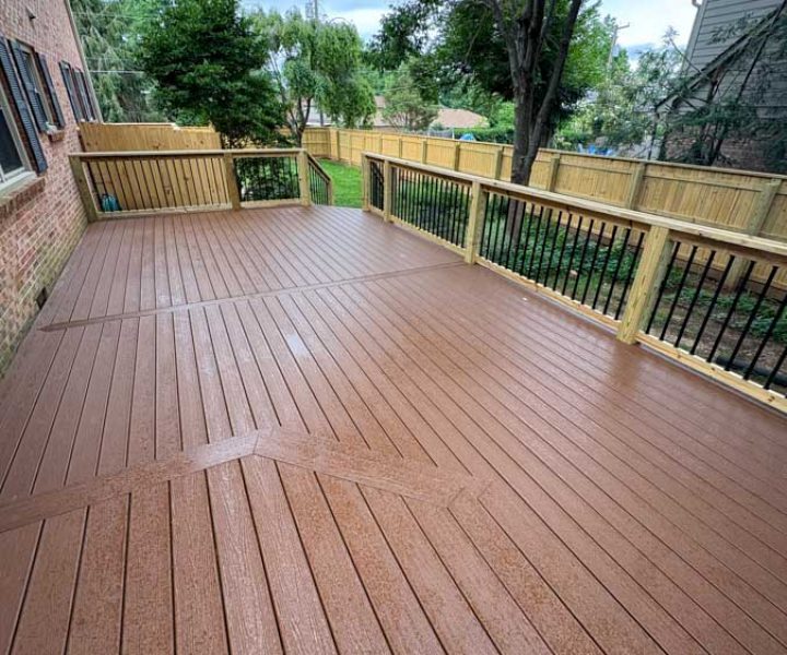 A spacious, newly built deck with rich brown composite flooring arranged in a geometric pattern, framed by natural wood railings with black balusters, set against a backdrop of trees and neighboring homes.